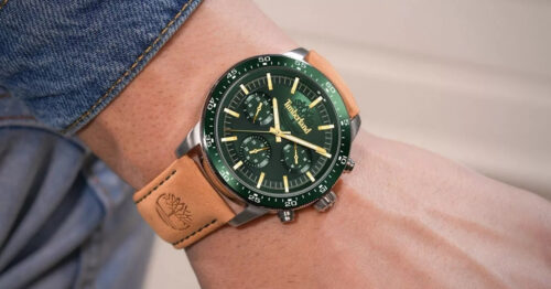 Green dial chronograph watch with a tan leather strap on a wrist, denim sleeve in the background.