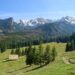 Snow-capped mountains overlook a green valley with pine trees and a wooden cabin in the foreground under a clear blue sky, bright day