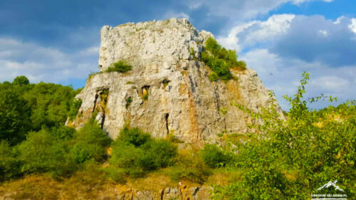 Large rocky limestone hill rising above green shrubland under a partly cloudy blue sky.
