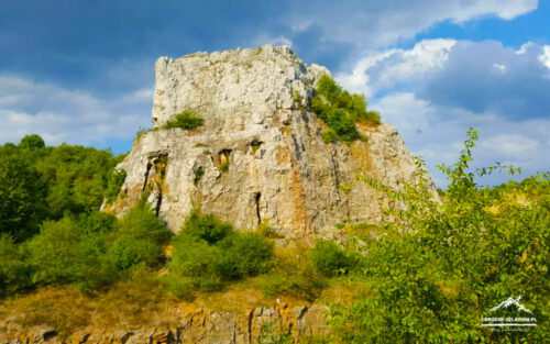 Large rocky limestone hill rising above green shrubland under a partly cloudy blue sky.