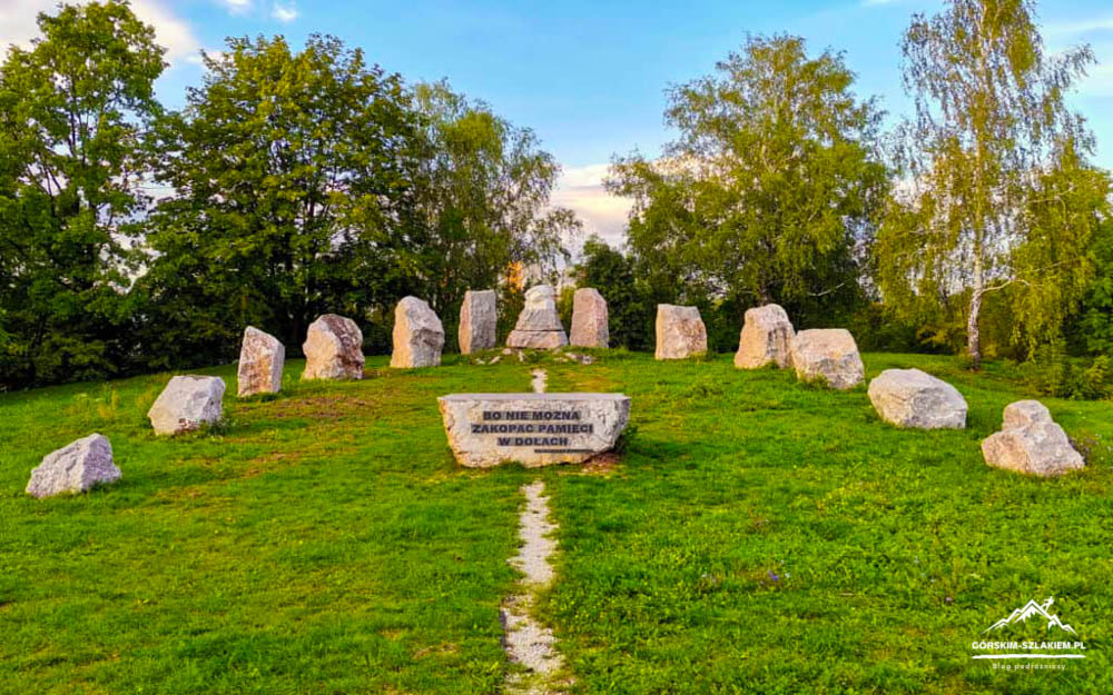 Circle of large standing stones on a grassy hill, with a central inscribed stone and surrounding trees under a blue sky (memorial site).