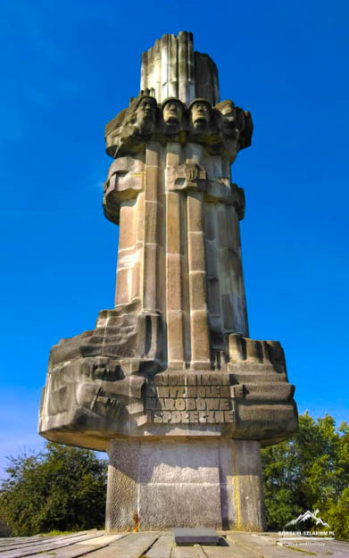 Stone monument: tall column crowned with five sculpted heads, with carved reliefs and an inscription at the base, set on a square pedestal against a blue sky.