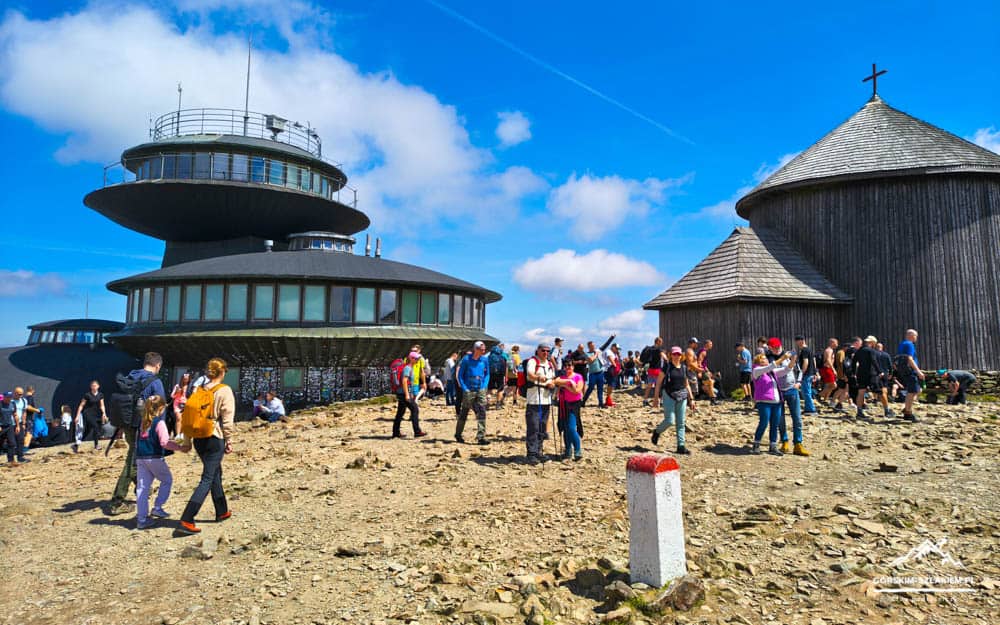 Group of hikers on a rocky hill near a circular modern building and a wooden church under a bright blue sky in daytime.