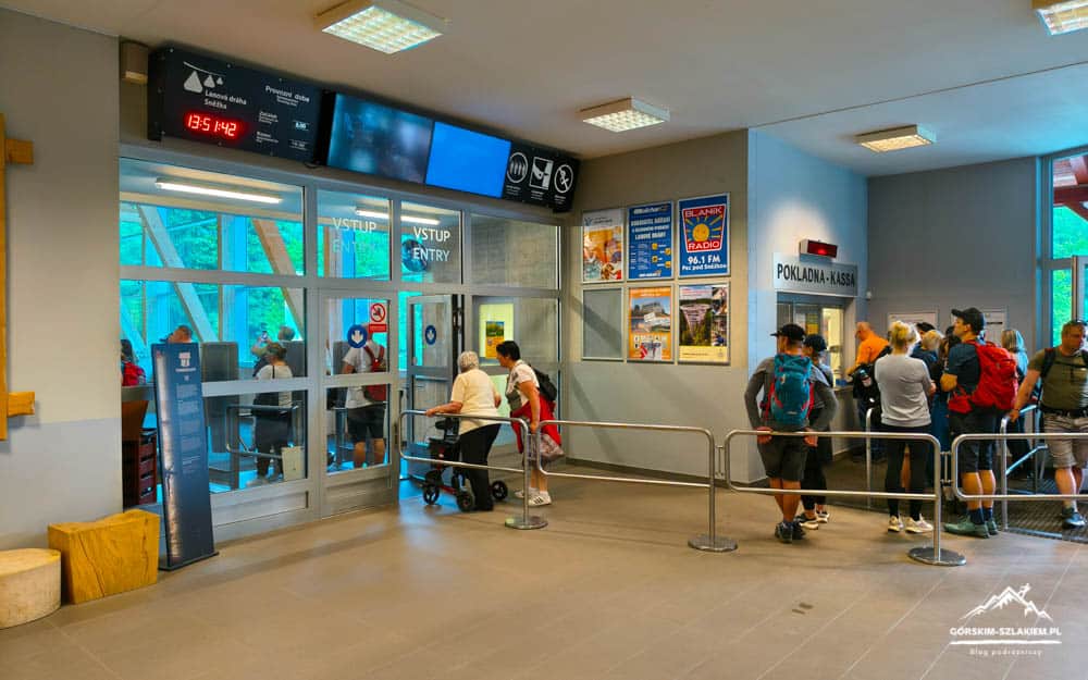 Indoor ticket hall with a line of people at a checkout counter behind glass doors labeled Pokladna-Kassa, posters on the wall, and a stroller nearby.