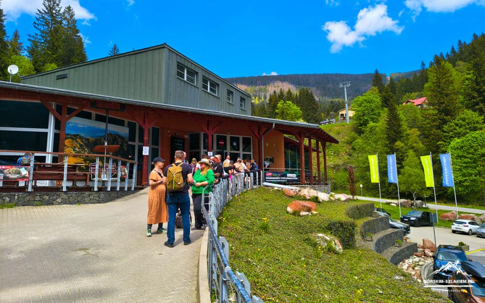 Group of visitors queuing outside a mountain lodge with a wooden veranda and a forested hillside in the background.