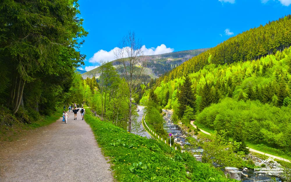 People walk along a dirt path through a green mountain valley with a river to the right.