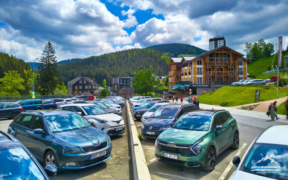 Busy mountain village parking lot filled with cars, a wooden hotel/café building in the background, and forested hills beyond.