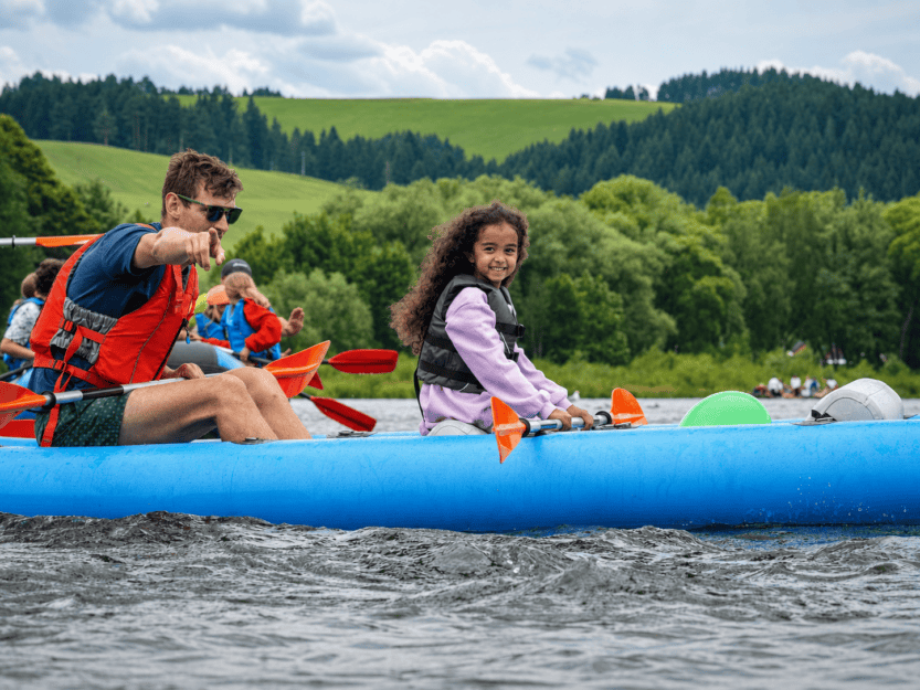 Two people in a blue inflatable raft on a lake, wearing life jackets and paddling, with the man pointing toward something ahead.