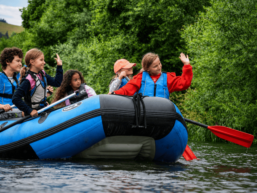 Group of kids in life jackets on a blue inflatable raft, waving and paddling on a river surrounded by green trees on a sunny day.
