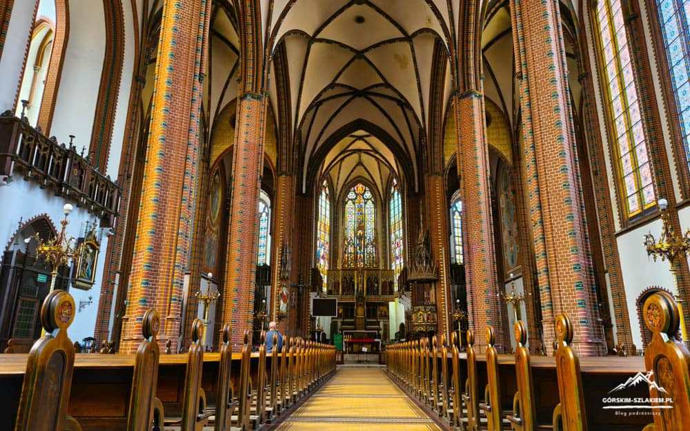 Gothic church interior with tall brick columns, ribbed vaults, and colorful stained-glass windows along a central aisle toward the altar.