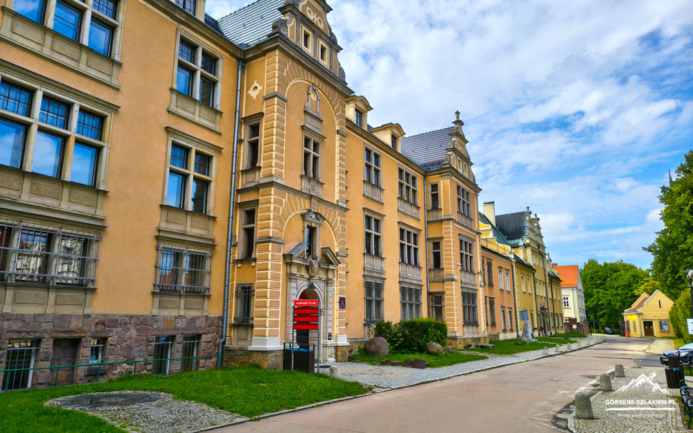 Row of yellow ornate historic buildings along a cobblestone street under a bright blue sky with green trees nearby.