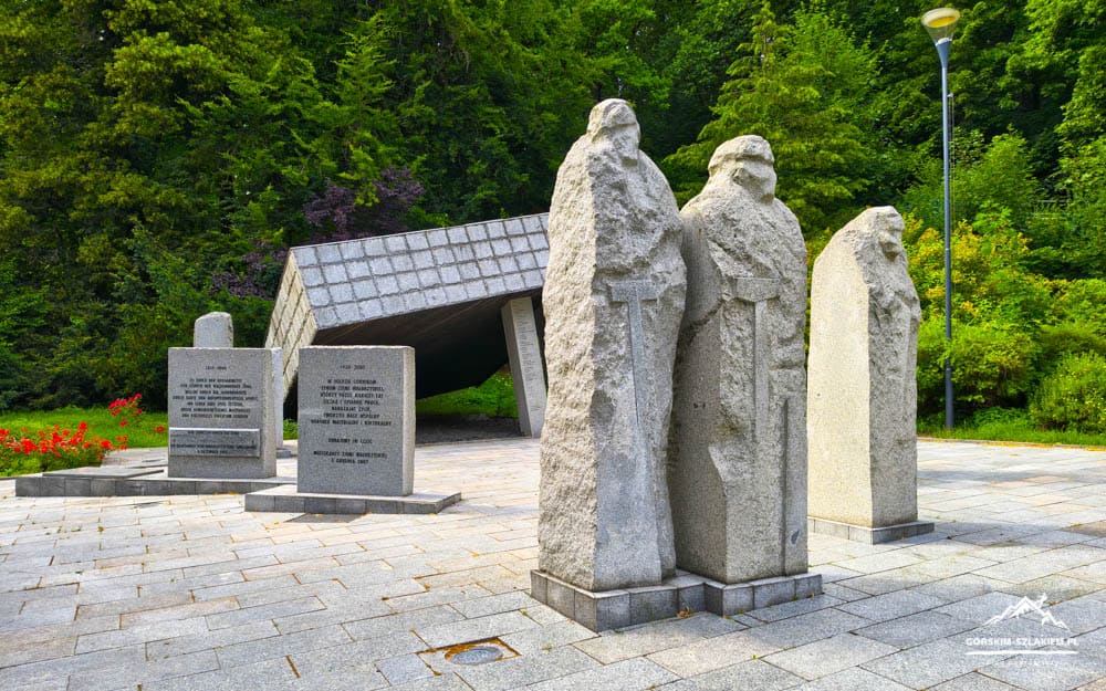Stone statues forming a group in a memorial plaza with plaques and a sloped stone structure, surrounded by green trees.