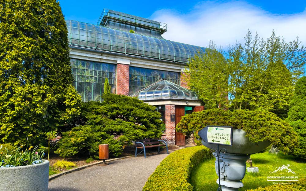Glass-walled greenhouse with brick entry, surrounded by lush shrubs and trees; sign reads Wejście / Entrance near a stone pedestal planter.