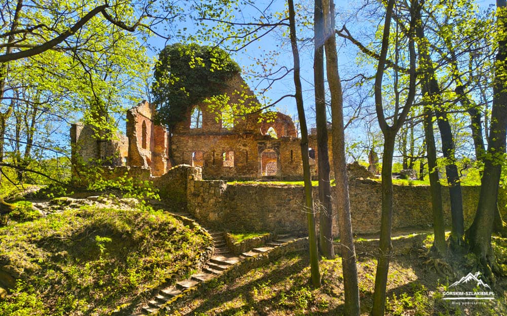 Ruins of a brick building atop a stone wall in a sunlit forest with a stone path nearby.