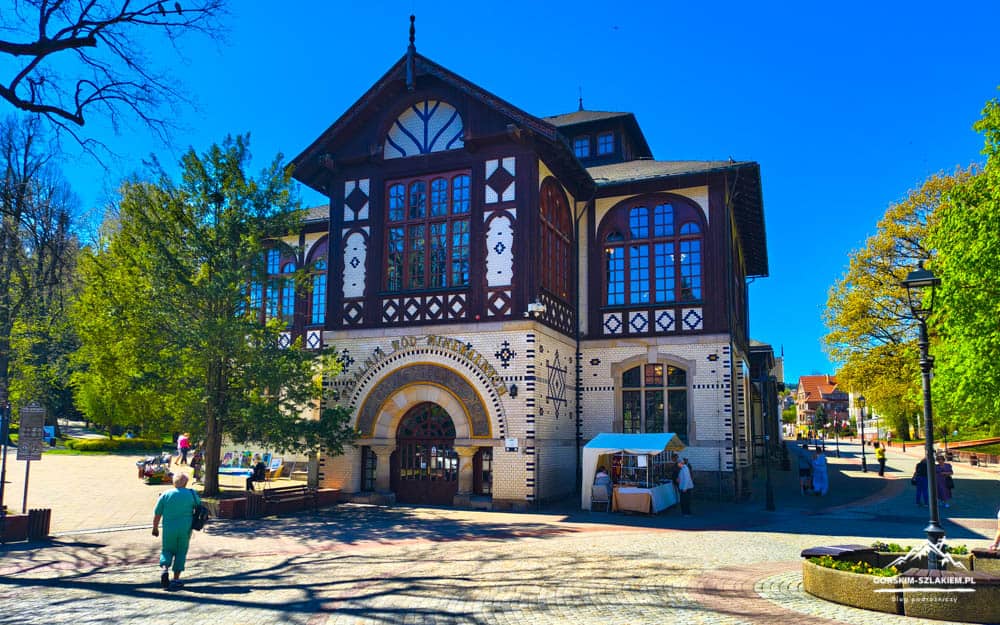Historic brick-and-timber building with arched windows in a sunny park plaza; people stroll and a market stall sits near the entrance.