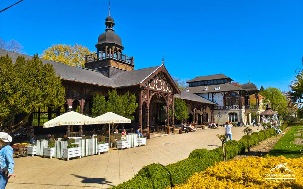 Sunny plaza with a wooden arcade cafe, outdoor seating under white umbrellas, and potted plants in front.