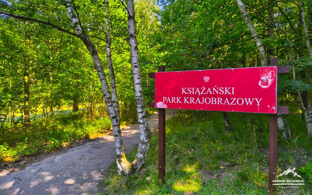 Red park sign reading 'Książąński Park Krajobrazowy' along a forest path