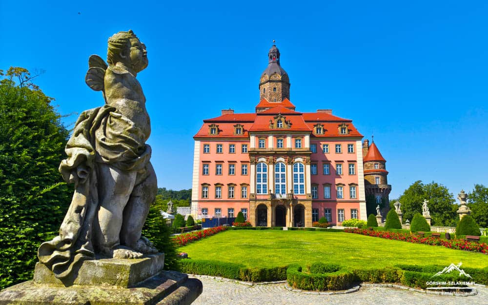Pink castle with red roof and central tower, framed by a stone statue and a manicured lawn on a bright day.