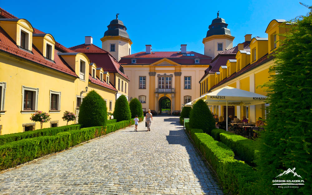 Sunlit courtyard of a yellow historic building with a cobblestone path, trimmed hedges, and an outdoor cafe on the right.