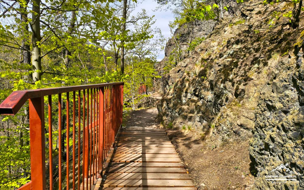 Wooden boardwalk with a red railing runs along a rocky cliff edge through a green forest.