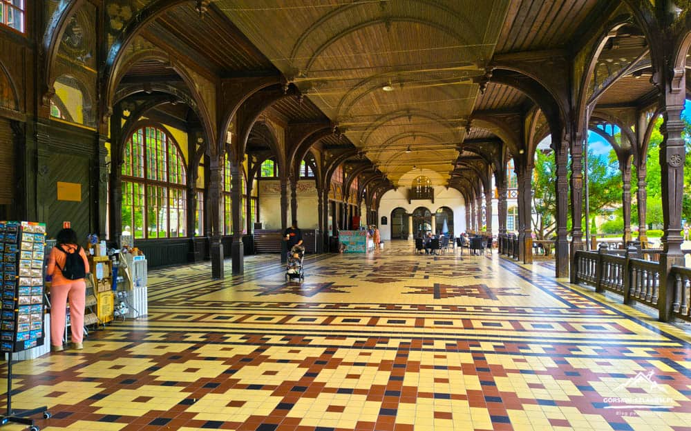 Historic arcade with dark wooden arches, a colorful tiled floor, and people browsing stalls on the left and trees visible on the right.