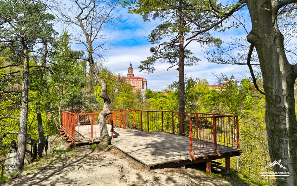 Wooden viewing deck with red railings in a forest, overlooking a distant city skyline with a domed building.