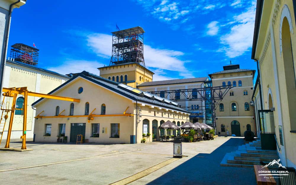 Sunlit courtyard of a historic industrial complex with pale yellow buildings, tall scaffolding towers, and outdoor cafe seating under umbrellas.