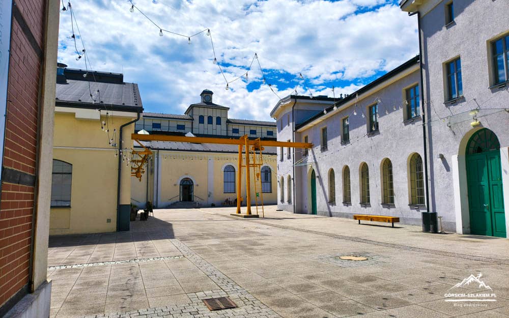 Sunny courtyard between pastel-colored buildings with string lights and an orange overhead gantry in the center, blue sky above.