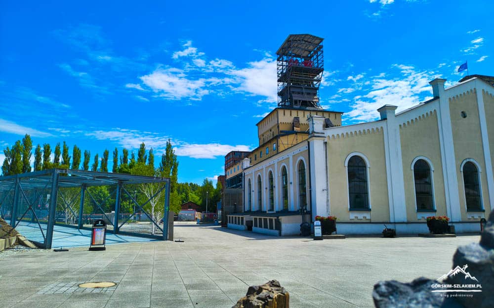 Historic building with a tall wooden observation tower, arched windows, and a glass-covered entrance on a sunny day in a paved plaza with trees in the background