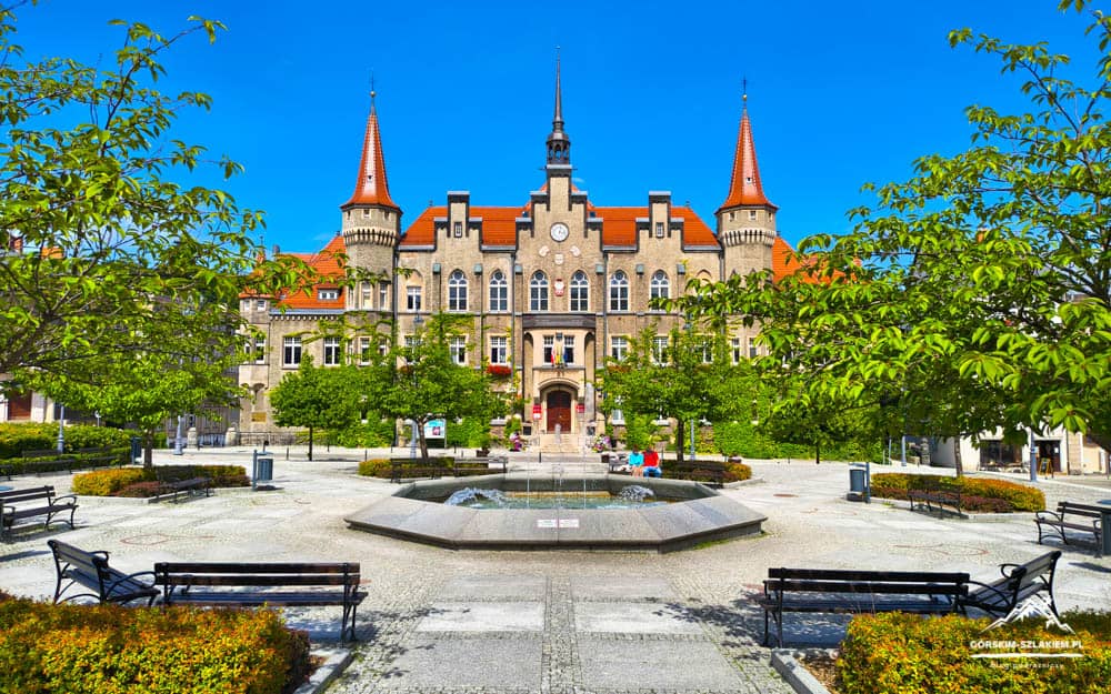 Historic beige brick town hall with red roofs and spires, flanked by trees, in a sunny town square with a central fountain.