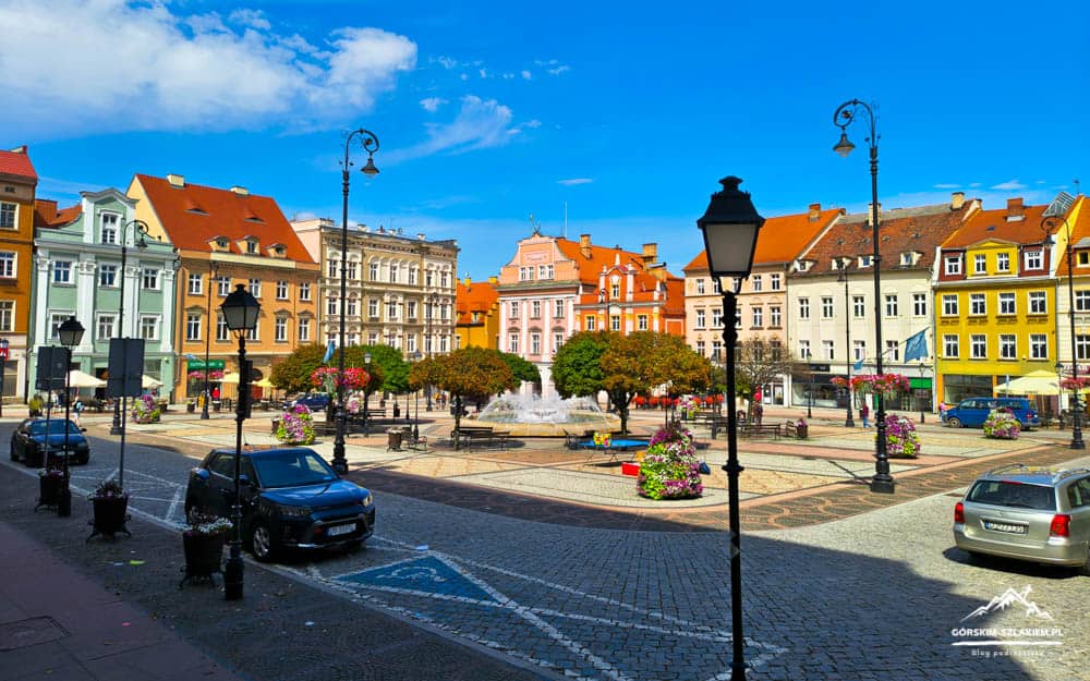 Colorful European town square with a central fountain, pastel buildings, benches, and parked cars under a bright blue sky.