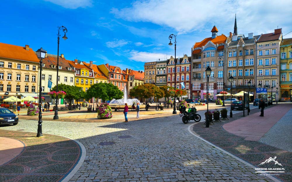 Colorful town square with a central fountain, outdoor seating, and pastel buildings under a bright blue sky.