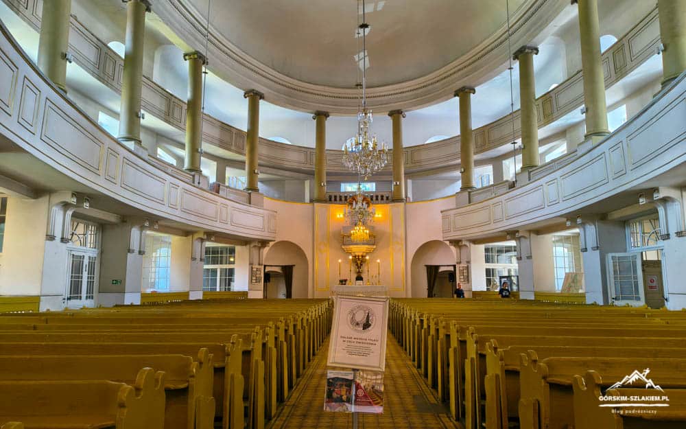 Interior of a grand circular church with balconies, columns, chandeliers, and rows of wooden pews facing an ornate altar area.