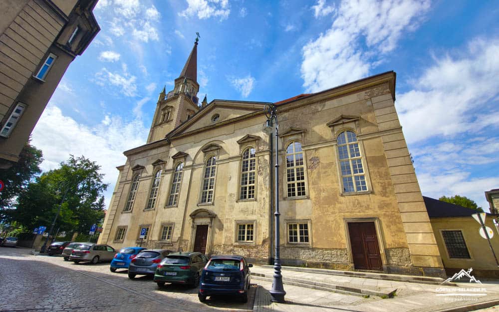 Historic church or town hall building on a sunny cobblestone street with parked cars and a church steeple against a blue sky.