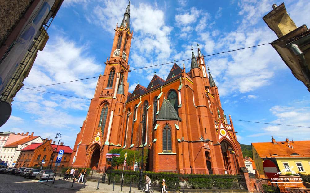 Red brick Gothic church with a tall clock tower in a sunny town square; pedestrians and parked cars nearby.