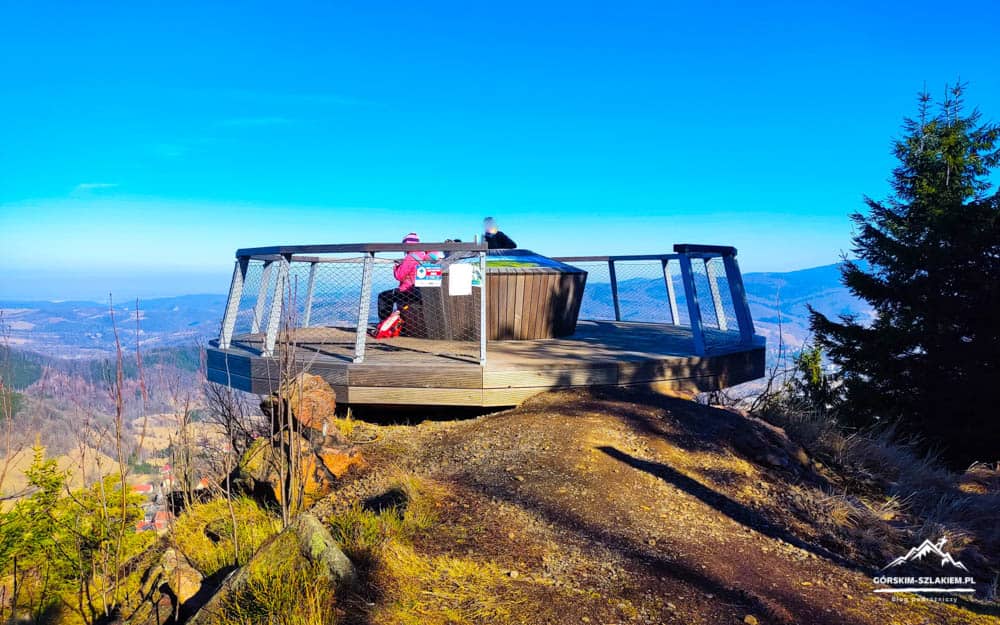 Wooden viewing platform with metal railings sits on a rocky hilltop, offering a panoramic valley view under a bright blue sky, with two people sitting at a bench.