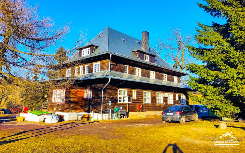 Two-story wooden house with a steep blue roof, white-framed windows, and surrounding trees on a sunny day, with a car parked outside.