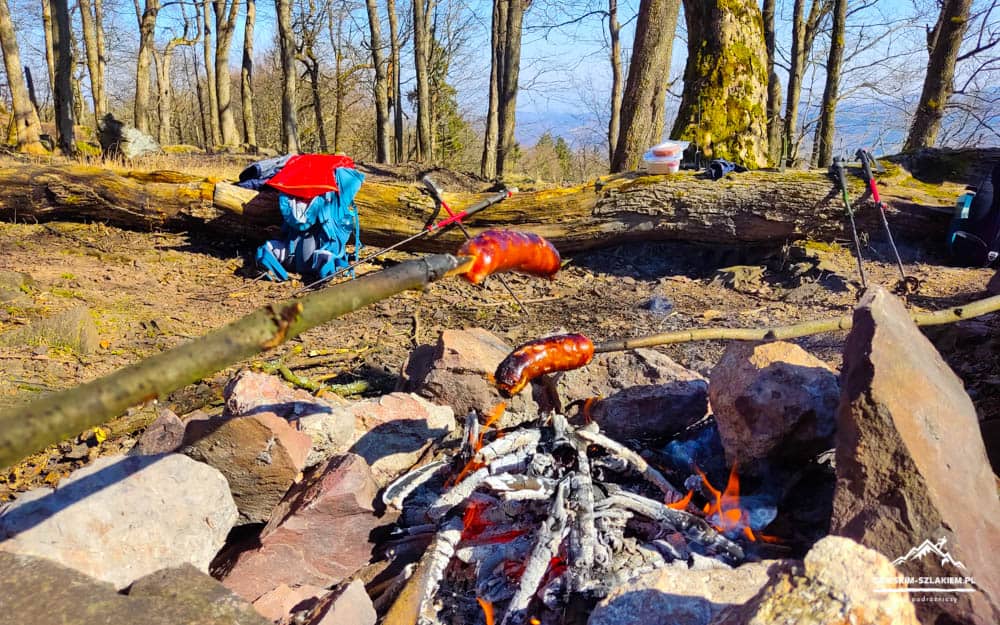 Campfire with sausages roasting over rocks in a wooded campsite, with backpacks and gear in the background.