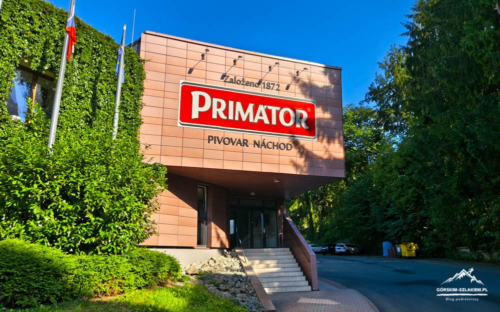 Exterior of Primator beer brewery building with a large red Primator sign and 'Pivovar Náchod' lettering, stairs to the entrance, surrounded by greenery on the left.