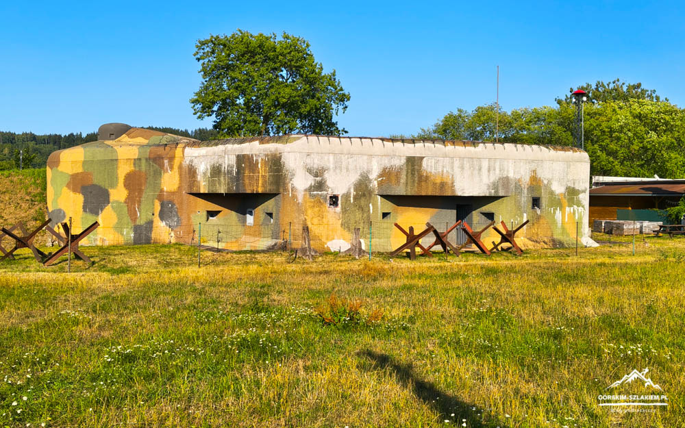 Concrete military bunker with camouflage painting in greens, browns, and yellows sits in a grassy field under a bright blue sky, with wooden X-shaped anti-tank barriers lined in front.