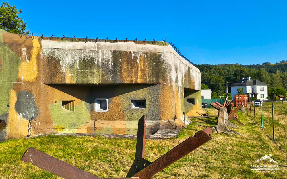 Abandoned concrete bunker with barbed wire on top, rusted metal structures in a grassy field under a bright blue sky.