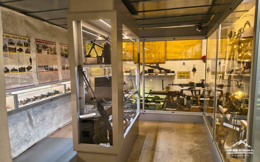 Interior of a weapons museum with glass display cases housing rifles and helmets along a corridor of exhibits.