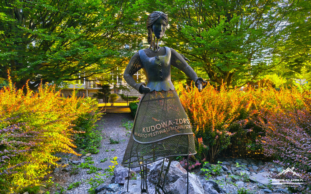 Metal statue of a woman in a long dress standing in a garden, with orange shrubs and green trees in the background; dress has text referencing Kudowa-Zdrój and a festival.