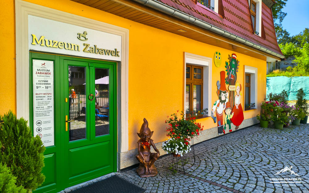 Bright yellow toy museum building with a green door, colorful mural, and cobblestone courtyard with plants.