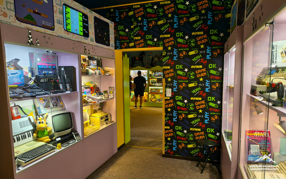 Retro gaming shop interior with glass display cases housing vintage computers and consoles, pink shelves, and a patterned wall backdrop.