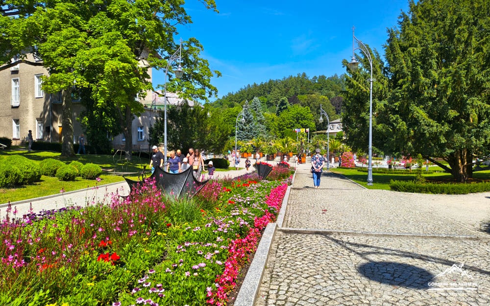 Sunlit park with colorful flower beds along a cobblestone path and people strolling under green trees.