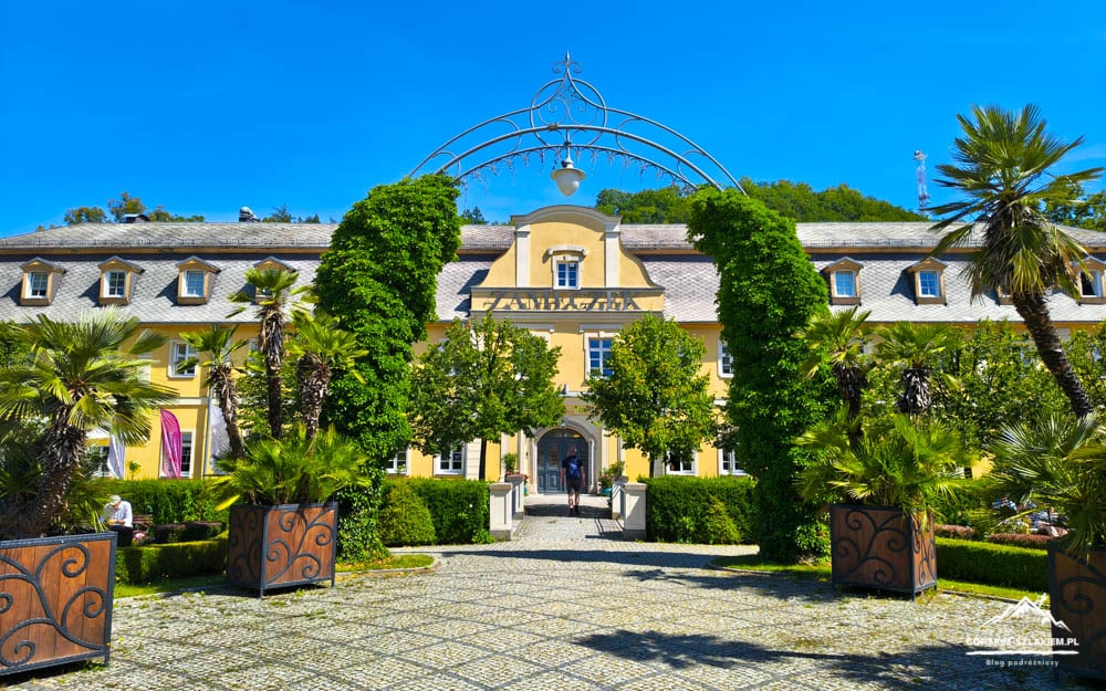 Yellow manor with an arched metal centerpiece, framed by greenery and palm trees, leading to a cobblestone plaza.