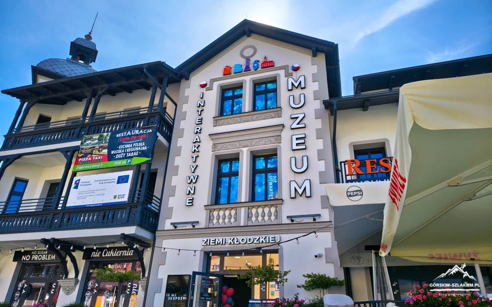 Facade of a Polish museum building with vertical 'MUZEUM' and 'INTERAKTYWNE' signs under a clear blue sky