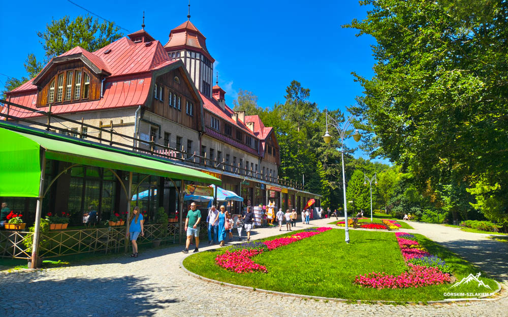 Historic red-roofed building with a green awning along a cobblestone street; people stroll in a sunny park with colorful flowerbeds nearby.