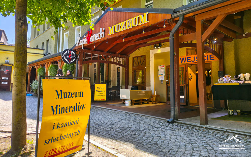 Entrance to a mineral museum with a yellow sign and wooden awning; cobblestone street and people nearby.